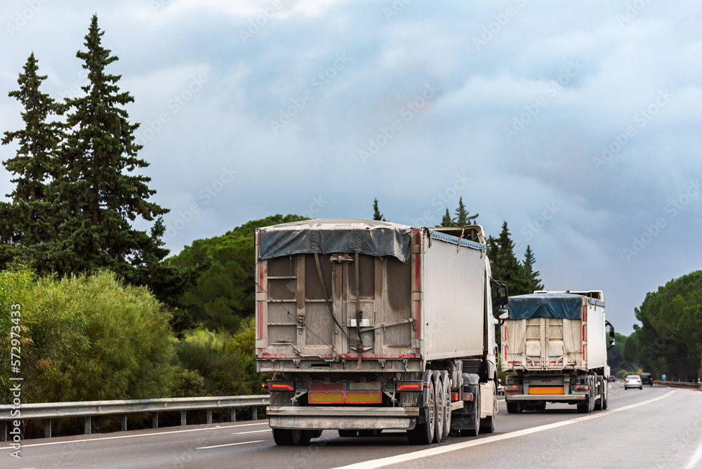 Trucks with tarpaulin tippers for bulk transport driving on a highway, rear view. Trucks with tarpaulin tippers for bulk transport driving on a highway, rear view.