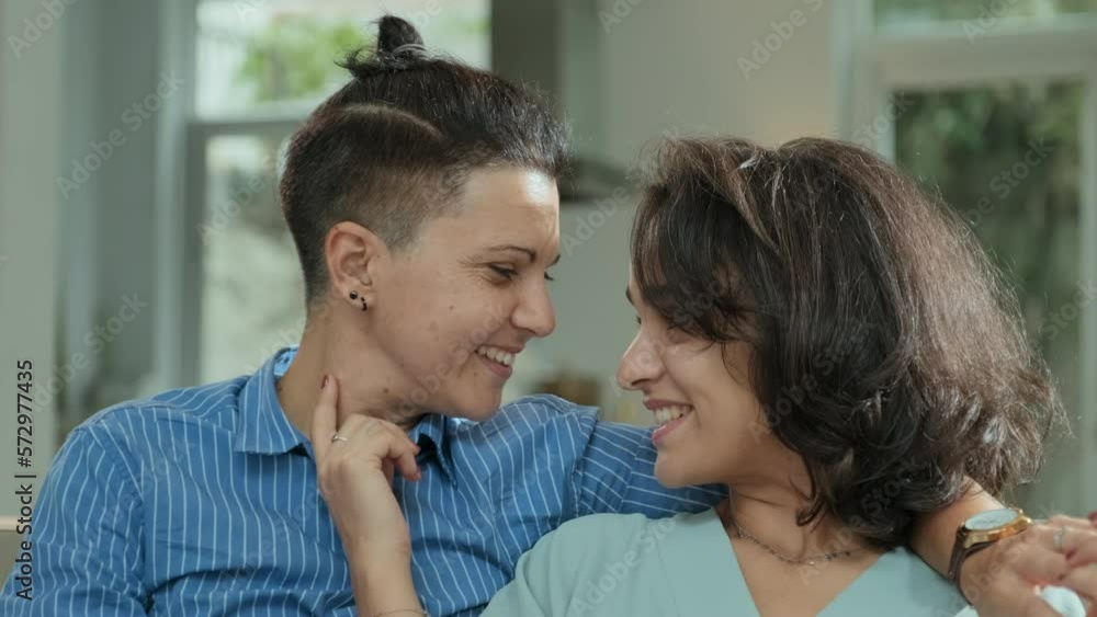 Tilt up shot of young Caucasian romantic lesbian couple holding hands and looking at each other with tenderness sitting on couch in living room together chatting and laughing Tilt up shot of young Caucasian romantic lesbian couple holding hands and looking at each other with tenderness sitting on couch in living room together chatting and laughing