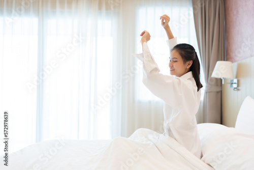 Asian women wake up in the morning In the bedroom of the house, the surrounding environment resembled a hotel. She smiled with freshness and relief. have good health wearing long-sleeved white pajamas