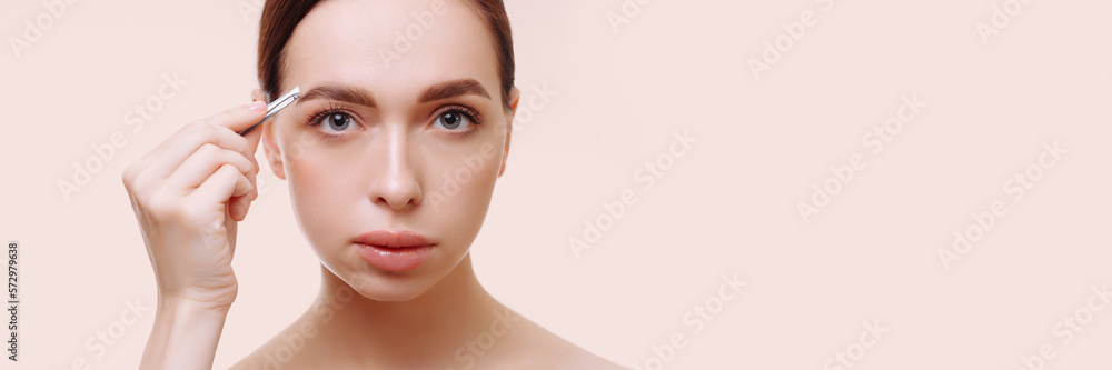 Banner portrait of an attractive young brunette woman plucking her eyebrows against a white background. Holds tweezers in hand. The concept of eyebrow care, cosmetic procedures.