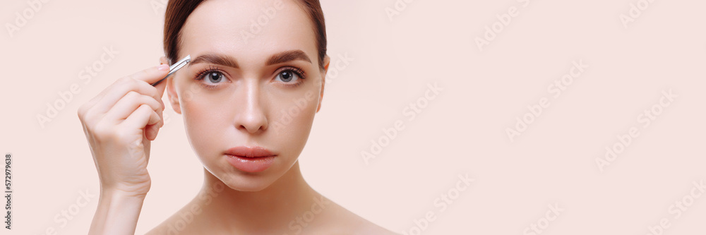 Banner portrait of an attractive young brunette woman plucking her eyebrows against a white background. Holds tweezers in hand. The concept of eyebrow care, cosmetic procedures. Banner portrait of an attractive young brunette woman plucking her eyebrows against a white background. Holds tweezers in hand. The concept of eyebrow care, cosmetic procedures.