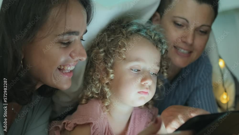 Two smiling Caucasian women in love and their cute little daughter lying under soft cozy blanket together at night using digital tablet Two smiling Caucasian women in love and their cute little daughter lying under soft cozy blanket together at night using digital tablet