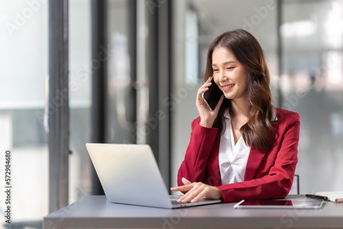 Cheerful young business woman talking on phone working in modern office. Happy positive Asian businesswoman company manager wearing suit making call on cellphone sitting at workplace
