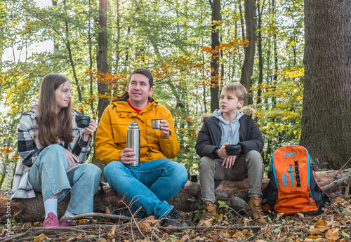 Father and children resting in nature, drinking hot tea a thermos in sunny autumn day.
