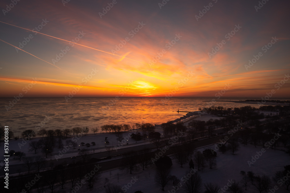 Aerial view of a colorful yellow orange and pink sunrise over Lake Michigan in winter with pieces of ice reflecting the orange glow on the water and snow on the ground between trees.