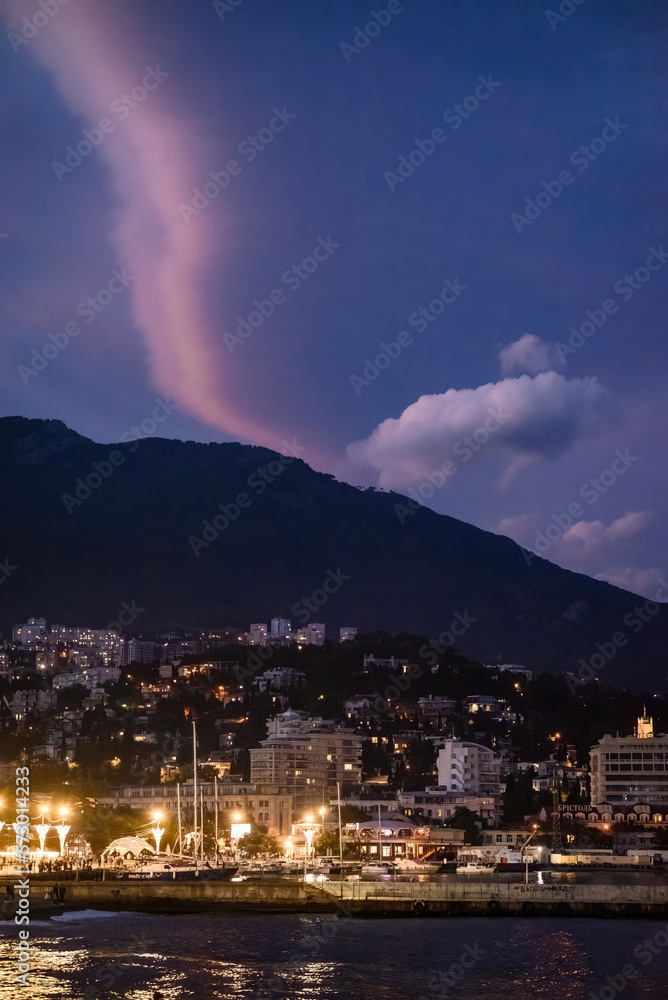Fototapeta premium Town On The Coast At Night. A coastal town with buildings and a harbor is nestled at the base of a dark mountain under a dramatic purple and dark blue sky.