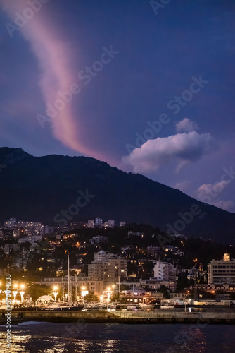 View of illuminated city on hill against backdrop of mountains and sky with clouds at dusk © EdvardPanov