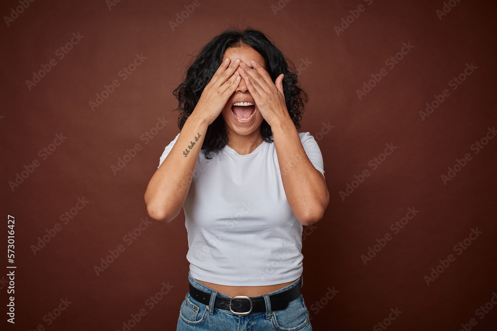 Young colombian curly hair woman isolated on brown background covers eyes with hands, smiles broadly waiting for a surprise.