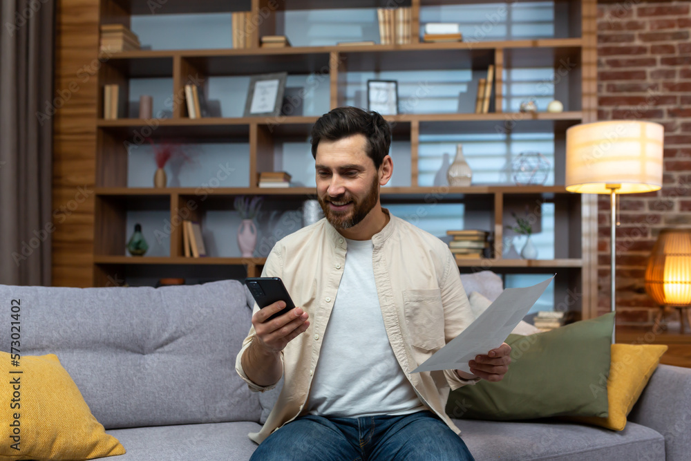 Cheerful and smiling man at home paying bills and checks, using banking app on phone sitting on sofa in living room holding bills.