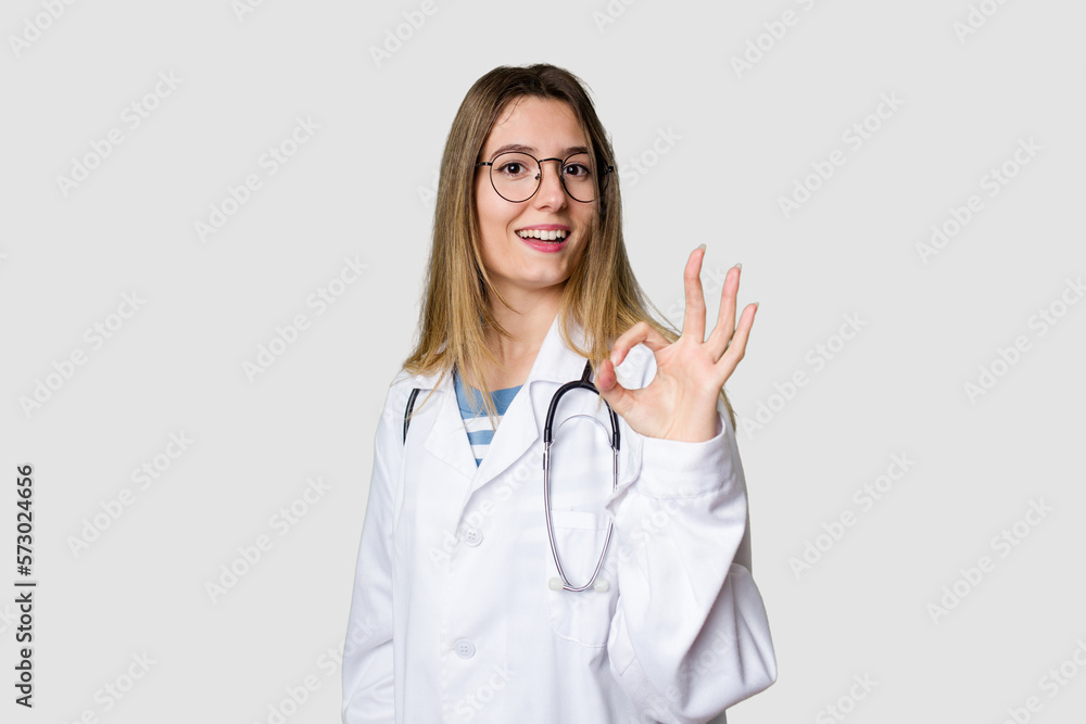 Compassionate female physician with a stethoscope around her neck, ready to diagnose and care for her patients in her signature white coat cheerful and confident showing ok gesture. Compassionate female physician with a stethoscope around her neck, ready to diagnose and care for her patients in her signature white coat cheerful and confident showing ok gesture.