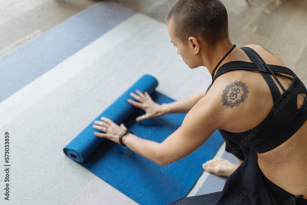 short haired tattooed woman unrolling yoga mat, preparing for yoga practice, behind view short haired tattooed woman unrolling yoga mat, preparing for yoga practice, behind view