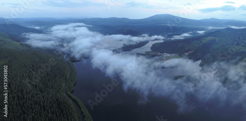 Aerial drone view above Oasa Lake and the wild spruce forests in Sureanu Mountains - at dusk. Fog and clouds are covering the water luster. Carpathia, Romania. photo