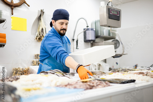 Photos A middle-aged fishmonger in an apron and a hat is about to catch a fish from the counter to put it in a box and finish work, selling food, small and medium-sized business