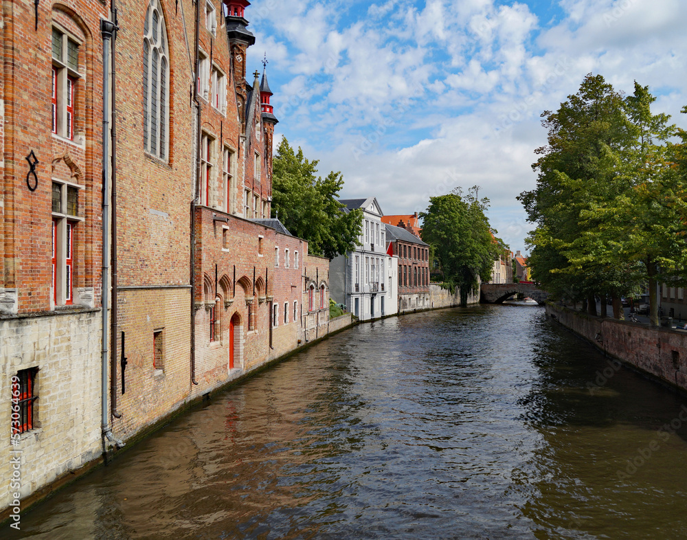 scenic sunlit canal in Bruges surrounded by the historic European architecture and lush green trees, Flanders, Belgium