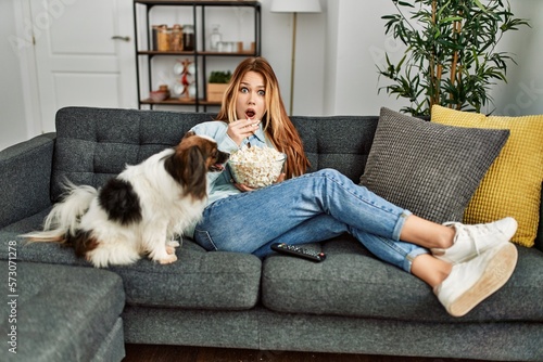 Young caucasian woman surprised watching movie sitting on sofa with dog at home