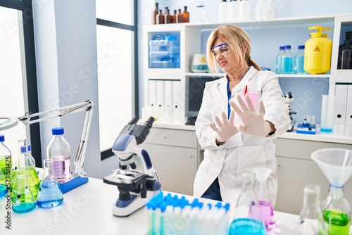 Middle age blonde woman working at scientist laboratory disgusted expression  displeased and fearful doing disgust face because aversion reaction.