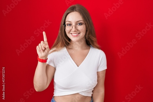 Young caucasian woman standing over red background showing and pointing up with finger number one while smiling confident and happy.