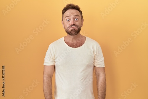Middle age man with beard standing over yellow background puffing cheeks with funny face. mouth inflated with air, crazy expression.