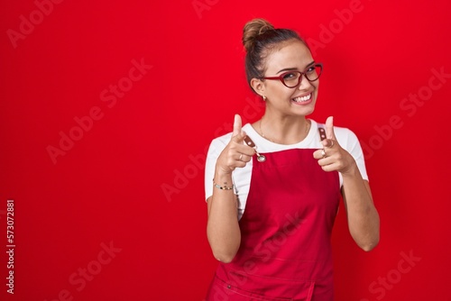 Fototapeta Young hispanic woman wearing waitress apron over red background pointing fingers to camera with happy and funny face