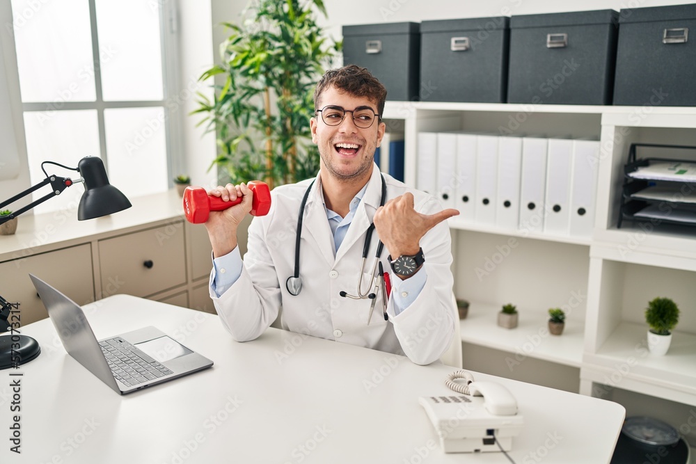 Young hispanic doctor man holding dumbbells pointing thumb up to the side smiling happy with open mouth