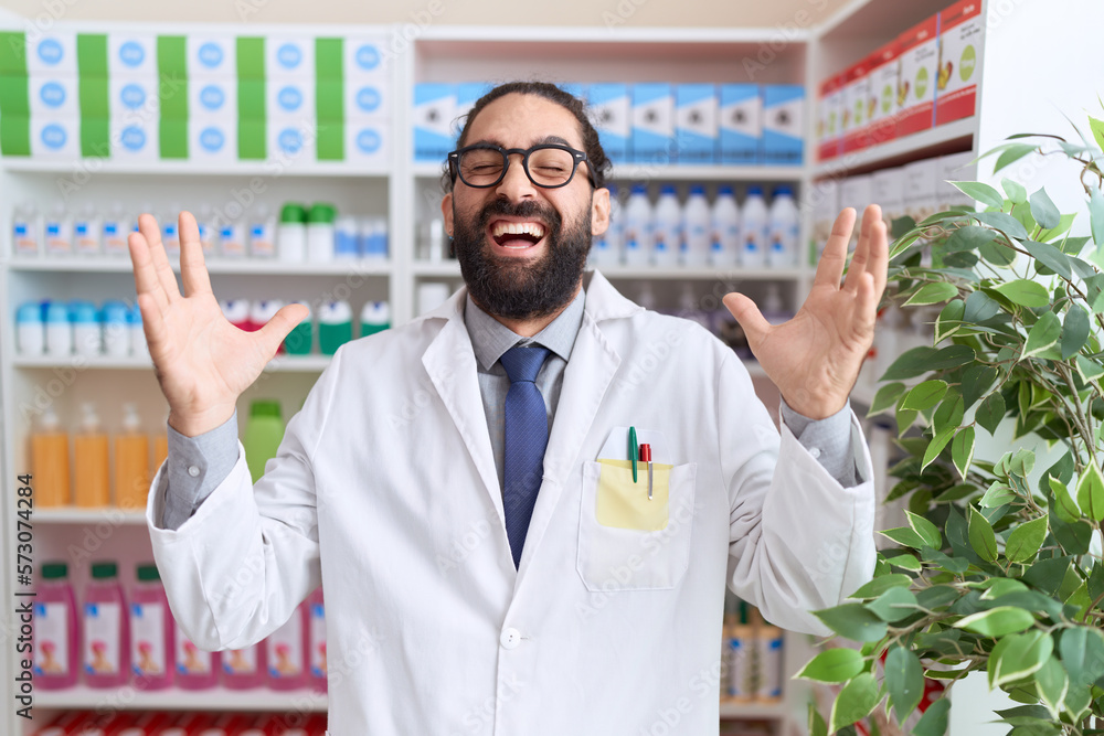 Hispanic man with beard working at pharmacy drugstore celebrating mad and crazy for success with arms raised and closed eyes screaming excited. winner concept
