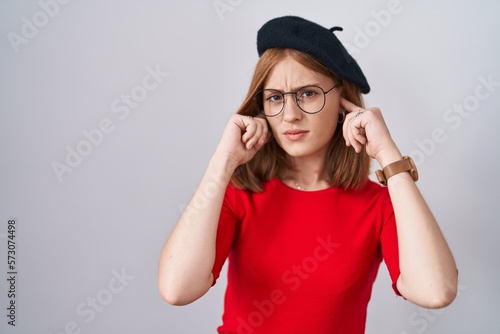 Young redhead woman standing wearing glasses and beret covering ears with fingers with annoyed expression for the noise of loud music. deaf concept.