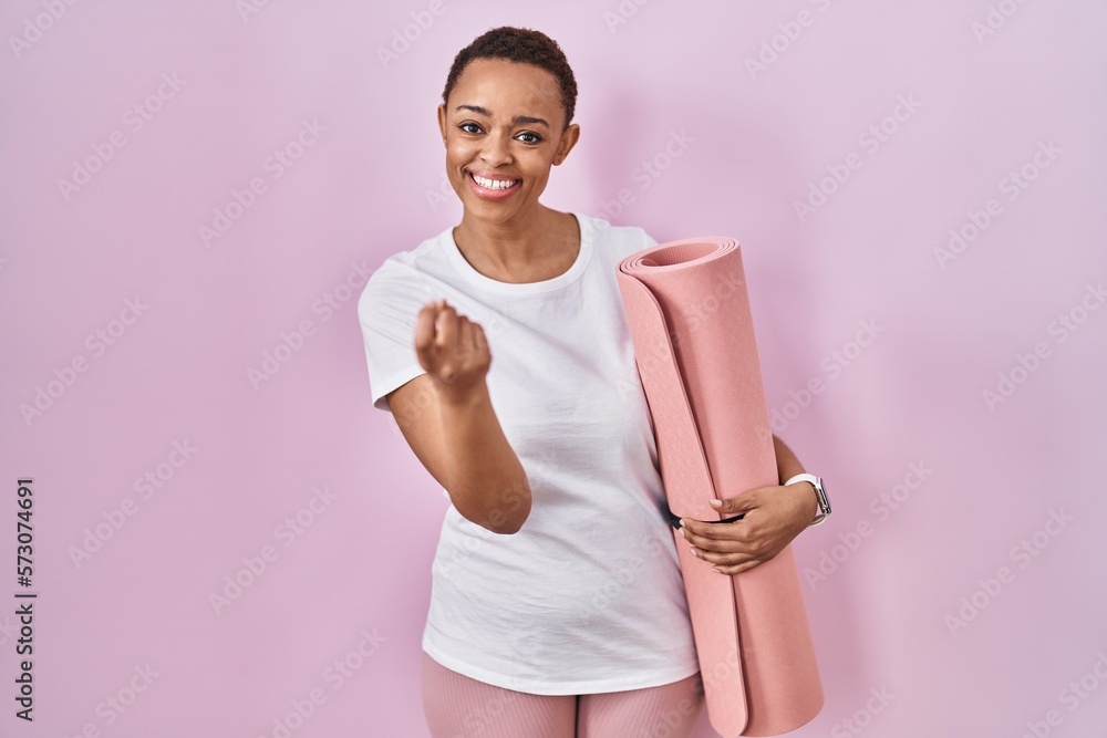 Beautiful african american woman holding yoga mat beckoning come here gesture with hand inviting welcoming happy and smiling