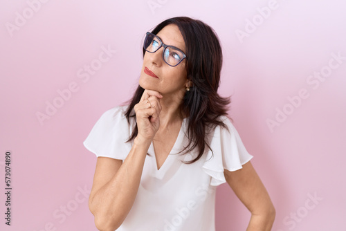 Middle age hispanic woman wearing casual white t shirt and glasses with hand on chin thinking about question, pensive expression. smiling with thoughtful face. doubt concept.