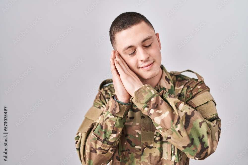 Young man wearing camouflage army uniform sleeping tired dreaming and posing with hands together while smiling with closed eyes.
