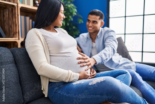 Young latin couple expecting baby touching belly sitting on sofa at home