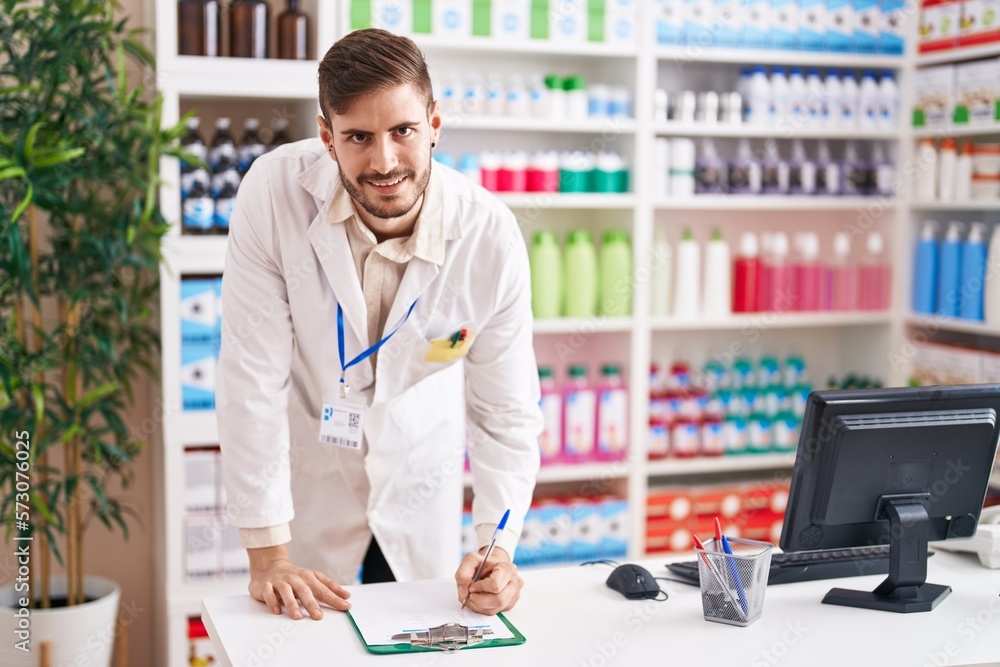 Young caucasian man pharmacist smiling confident writing on document at pharmacy