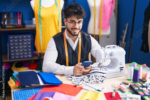 Young hispanic man tailor smiling confident using smartphone at sewing studio