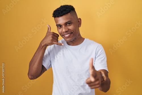 Young hispanic man standing over yellow background smiling doing talking on the telephone gesture and pointing to you. call me.