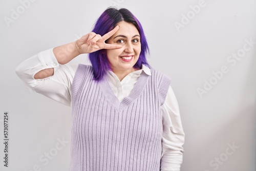 Plus size woman wit purple hair standing over white background doing peace symbol with fingers over face, smiling cheerful showing victory © Krakenimages.com