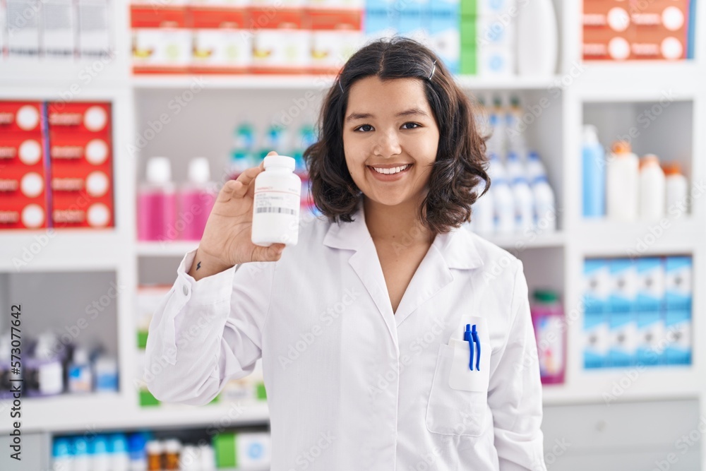 Young woman pharmacist smiling confident holding pills bottle at pharmacy