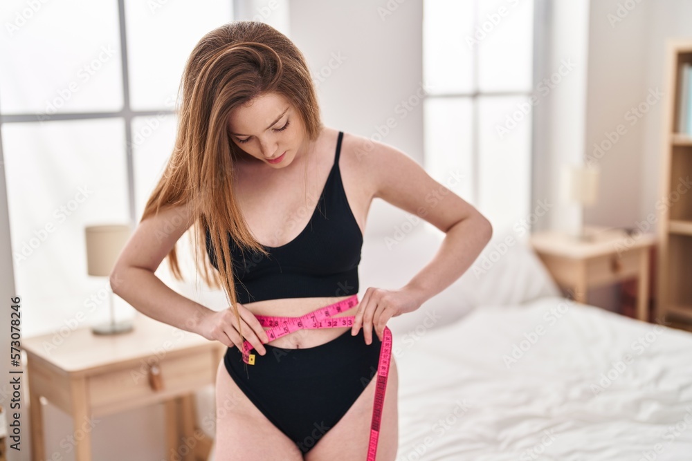 Young caucasian woman measuring waist at bedroom