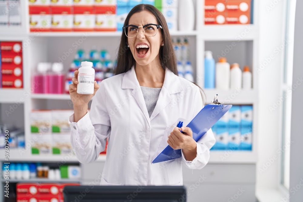 Young brunette woman working at pharmacy drugstore holding pills angry and mad screaming frustrated and furious, shouting with anger. rage and aggressive concept.