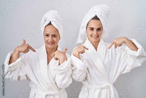 Middle age woman and daughter wearing white bathrobe and towel looking confident with smile on face, pointing oneself with fingers proud and happy.