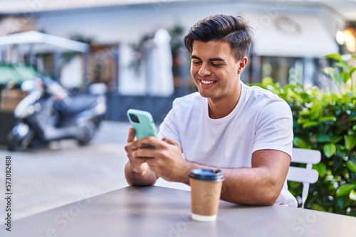 Young hispanic man using smartphone drinking coffee at coffee shop terrace