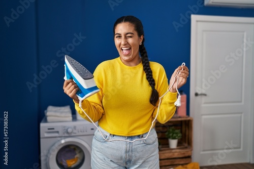 Young hispanic woman holding iron at laundry room winking looking at the camera with sexy expression, cheerful and happy face. © Krakenimages.com