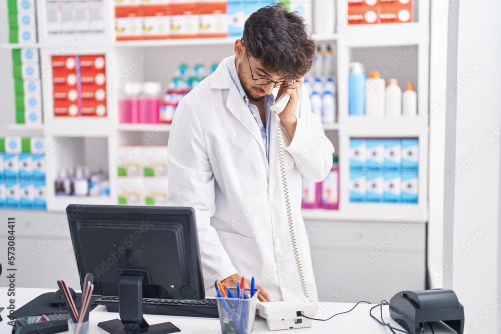 Young arab man pharmacist using computer talking on telephone at pharmacy