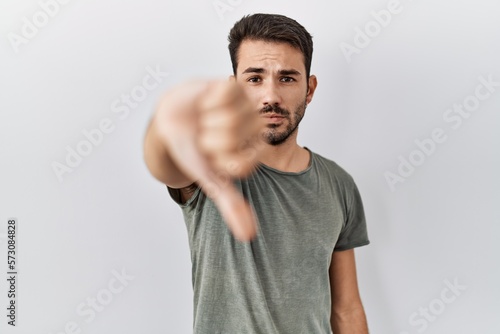 Young hispanic man with beard wearing casual t shirt over white background looking unhappy and angry showing rejection and negative with thumbs down gesture. bad expression.