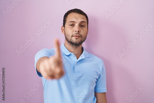 Hispanic man standing over pink background pointing with finger up and angry expression, showing no gesture