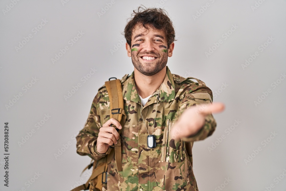 Hispanic young man wearing camouflage army uniform smiling cheerful offering palm hand giving assistance and acceptance. Hispanic young man wearing camouflage army uniform smiling cheerful offering palm hand giving assistance and acceptance.