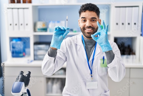 Hispanic man with beard working at scientist laboratory holding syringe doing ok sign with fingers  smiling friendly gesturing excellent symbol