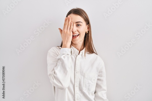 Young caucasian woman standing over isolated background covering one eye with hand, confident smile on face and surprise emotion.
