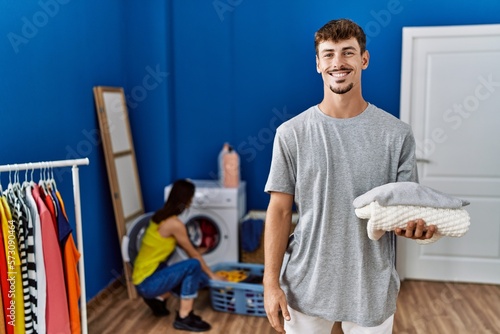 Young handsome man holding folded laundry looking positive and happy standing and smiling with a confident smile showing teeth