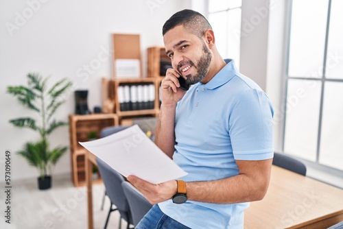Young latin man business worker talking on smartphone reading document at office