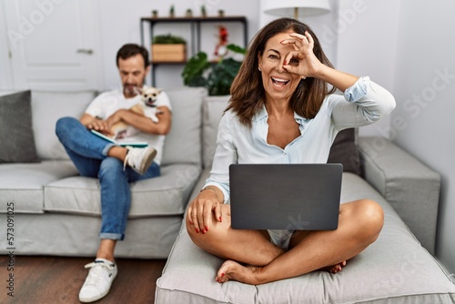 Hispanic middle age couple at home, woman using laptop doing ok gesture with hand smiling, eye looking through fingers with happy face.
