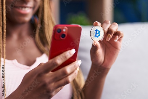 African american woman using smartphone holding bitcoin at home